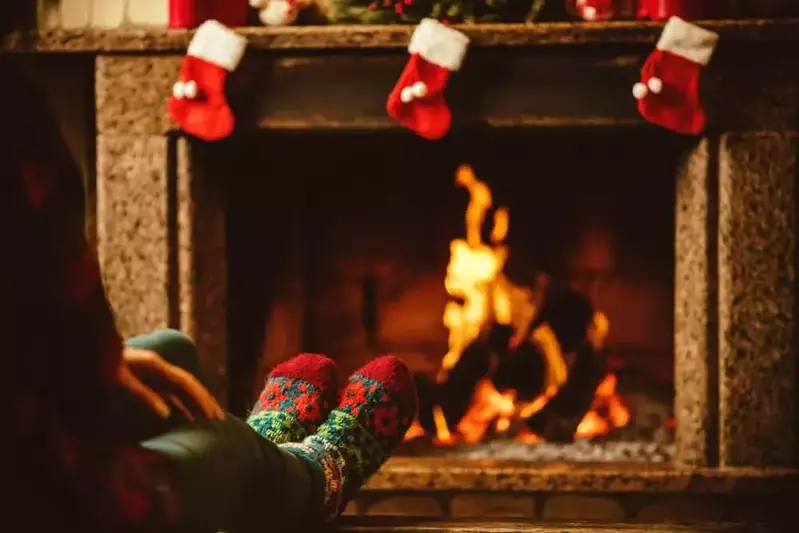 A woman warming her feet beside a fireplace in a cabin decorated for Christmas.