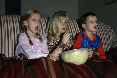 Three kids watching a movie, eating popcorn on the couch while sitting under a red blanket